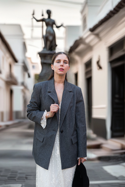 Frau in weißem Spitzenkleid und grauem Blazer in einer ruhigen Gasse mit Skulptur im Hintergrund. Sie trägt feinen Silberschmuck, die Szene wirkt elegant und urban.