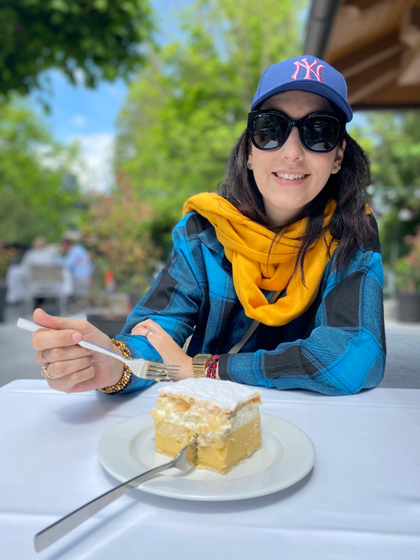 Carolina in camicia a scacchi blu e neri e sciarpa arancione e berretto blu, con una forchetta in mano sta per addentare una fetta della cremsnita, dolce sloveno a base di crema e panna