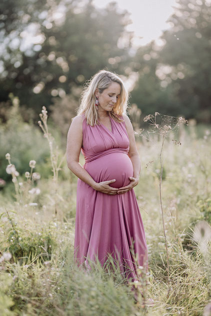 eine schwangere Frau in einem rosafarbenen Kleid steht auf einer Wiese und hält die Hände unter ihren Bauch
