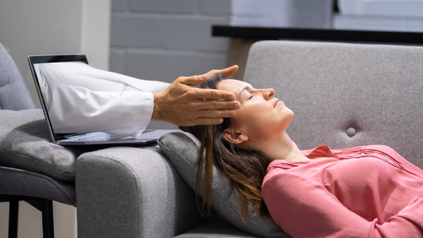 Fernbehandlung -Eine Frau entspannt auf einem Sofa während einer virtuellen Fernbehandlung, bei der die Hände eines Therapeuten aus dem Bildschirm eines Laptops herauskommen und sanft ihren Kopf berühren, was eine Online-Energietherapie darstellt.