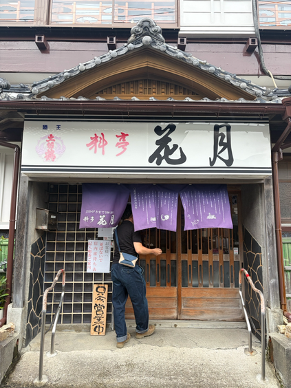 高知県の室戸にあるキンメ丼が有名な料亭花月の暖簾をくぐる黒いシャツとデニムを履いた男性。その男性は、斜めがけにぴったりな帆布バッグcaramelをかけいています
