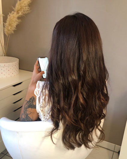 Cette photo représente une femme de dos qui regarde son smartphone et qui vient de recevoir une pose d'extensions de cheveux naturels chatains jusqu'en bas du dos.