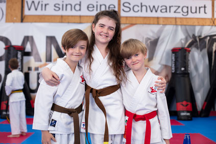 3 Kinder umarmen sich und sind fröhlich. Alle drei lachen und haben spaß beim Karate Training.