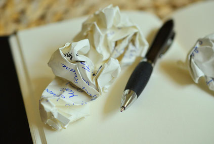 Auf einem Notizbuch liegen ein zerknülltes Blatt Papier und ein schwarzer Kugelschreiber. Auf dem Blatt stehen einzelne mit Kugelschreiber geschriebene Wörter. Der Fokus liegt auf dem Blatt und der Mine des Schreibers.
