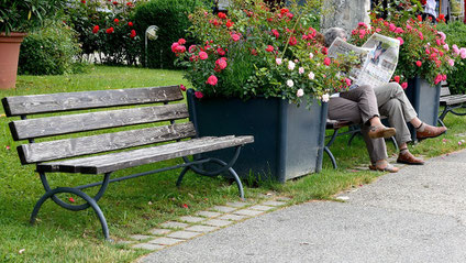 Zwei Parkbänke auf einem Rasen, dazwischen große Blumenkübel. Auf der rechten Bank, ziemlich weit in der rechten oberen Ecke, zwei Menschen, die aufgeschlagene Zeitungen halten, so dass man die Gesichter nicht sieht.