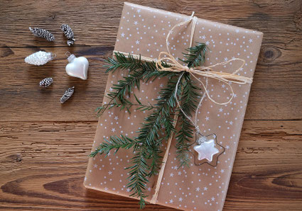 Ein Geschenk auf einem groben Holztisch. Vom Format könnte ein Buch drin sein. Es ist in grobes beiges Geschenkpapier mit weißen Sternen eingewickelt und mit einer Bastschnur umwickelt, darunter stecken ein Tannenzweig und ein sternförmiger Anhänger.