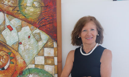 Margit Königsecker steht neben einem Gemälde, trägt ein schwarzes Kleid mit Perlenkette und sieht in die Kamera