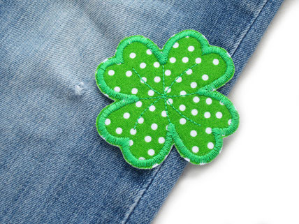 !B auf einer blauen Jeanshose liegt ein grüner Kleeblatt Patch zum aufbügeln, bereit um als Flicken das kleine Loch zu reparieren
