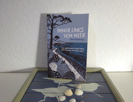 Das Buch  „Immer links vom Meer“ steht auf einem Bild aus Holzleisten, auf dem die Silhouetten von drei Strandvögeln in Weiß auf graublauem Grund zu sehen sind. Vor dem Buch liegen einige Muscheln.