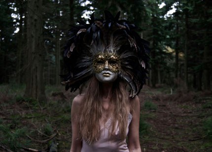 Eine Frau mit einer venezianischen Maske steht scheinbar deplatziert im Wald.