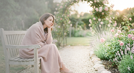 Femme pensive assise sur un banc de jardin, enveloppée dans un plaid rose poudré, illustrant la fatigue et le manque d'énergie – Accompagnement Sophrologie Haute-Loire.