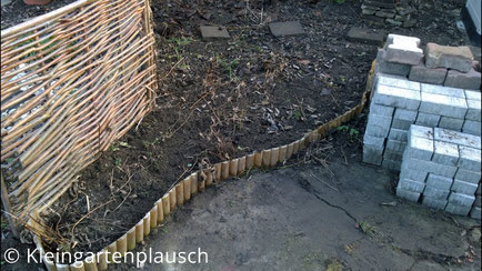 Im Vordergrund rechts gestapelte Pflastersteine, links ein Sichtschutzelement, davor kleine Holzpalisaden, die ein Beet vom Betonvorplatz vor der Hütte trennen