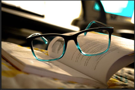 Auf einem aufgeschlagenen Buch liegt eine Brille. Der Rahmen ist oben schwarz, unten durchscheinend türkis. Im Hintergrund erkennt man ein Leuchten wie von einem Computermonitor.