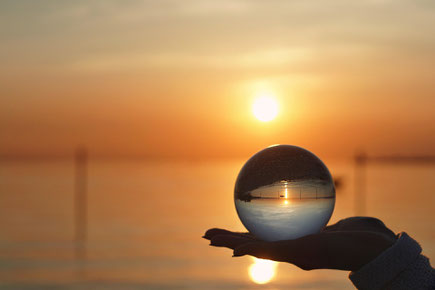 Eine Hand, die nur als Silhouette zu sehen ist, hält eine Glaskugel vor einem Sonnenuntergang am Wasser. Die Kugel zeigt den Sonnenuntergang über Kopf.