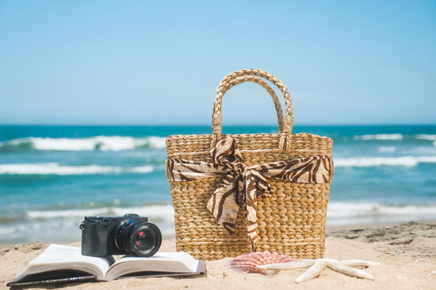 波しぶきを上げる海。砂浜に置かれた籐のバッグ。ページが開かれた本の上に置かれた一眼レフカメラ。