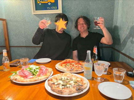 石川県金沢市にあるピザ屋サリーナにて、黒いシャツと黒い長袖シャツを着た男性二人がお酒を片手にもち笑顔でニコリ。そしてテーブルには生ハムが乗ったピザとマルゲリータ、サルシッチャのピザが３枚とレモネードが入った瓶、コップ、皿が２枚並んでいます。
