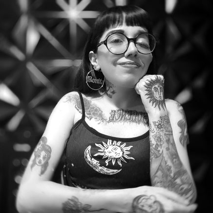 Dahlia de la Cerda trägt schwarze glatte Haare mit einem Pony, eine große Brille, große Creolen, ein Trägertop und viele Tattoos.