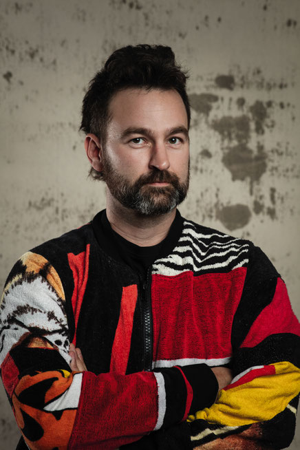 Das Foto zeigt den Autor Stephan Roiss mit Bart und einer bunten Jacken. Er hat die Arme vor der Brust verschränkt.