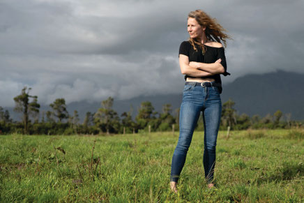 Auf dem Bild steht die Autorin Becky Manawatu  mit verschränkten Armen auf einer Wiese und die Haare wehen im Wind.