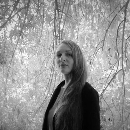 Johanna Malcher steht in einen Wald und schaut von oben herab in die Kamera. Sie trägt lange blonde Haare und einen schwarzen Blazer.