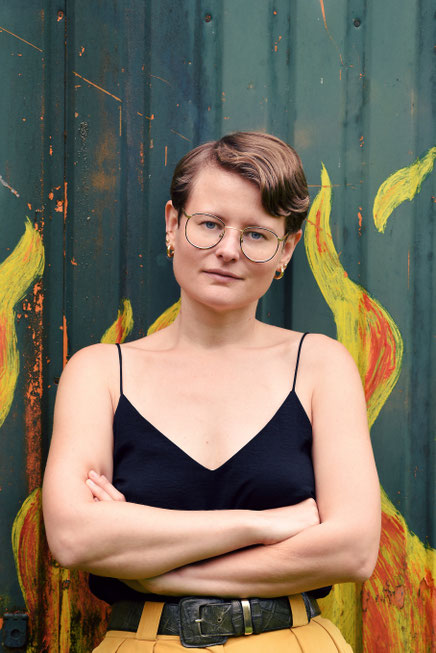 Das Bild zeigt die Autorin Thea Mengeler mit Spaghettishirt, Brille, kurzen Haaren und verschränkten Armen.