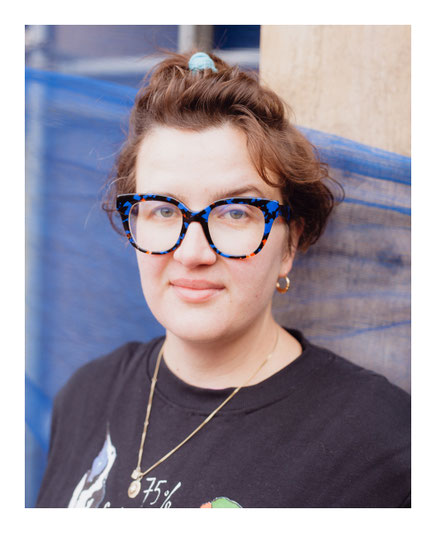 Das Bild zeigt die Autorin Katharina Volckmer mit lockigem Haar und bunter Brille, die eine AUgenbraue hochzieht.