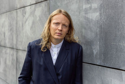 Das Bild zeigt den Autor Glenn Bech im Anzug und mit langen blonden Haaren an einer Wand lehnend.