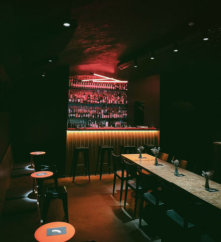 Die Bar Schacht in Siegen, gemütliche Atmosphäre, gedimmtes Licht, Tische und Stühle in der Mitte, rotes Licht strahlt auf die Theke mir vier Barhockern, großes Flaschenregal im Hintergrund der Theke