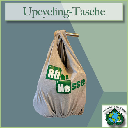 Graue Upcycling-Tasche aus einem Rhoihesse-T-Shirt, unten mit geknotetem Boden, hängend an einem Türgriff.