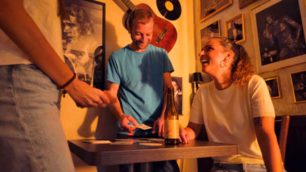 Drei Personen spielen den Escape Room Tödliche Musik Luzern. Auf dem Tisch stehen eine leere Flasche und Spielkarten. Ein Strategiespiel für Denker an Regentagen.