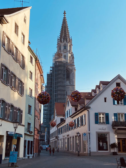 Blick auf den halb eingerüsteten Münsterturm vom Münster ULF in Konstanz. Blick in die Wessenbergstraße mit wunderschönen Altstadthäusern und großen Blumenkugeln, die über der Gasse hängen als Blumendeko. Teeladen links im roten Gebäude eröffnet 1.10.2026