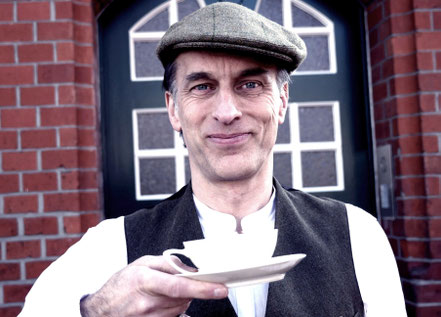 Thommi Baake steht lächelnd vor einer alten blauen Tür an einem Klinkerbau, hält eine Teetasse