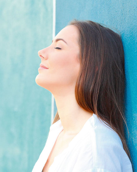 Frau lehnt versonnen an einer Wand und lässt sich die Sonne ins Gesicht scheinen.