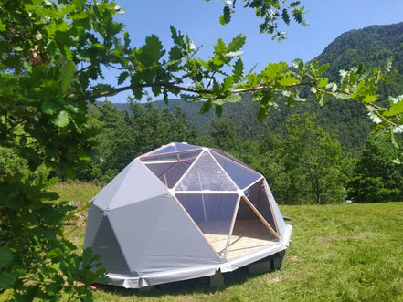 Dôme géodésique en toile grise sur pilotis, installé dans un pré avec vue sur les montagnes
