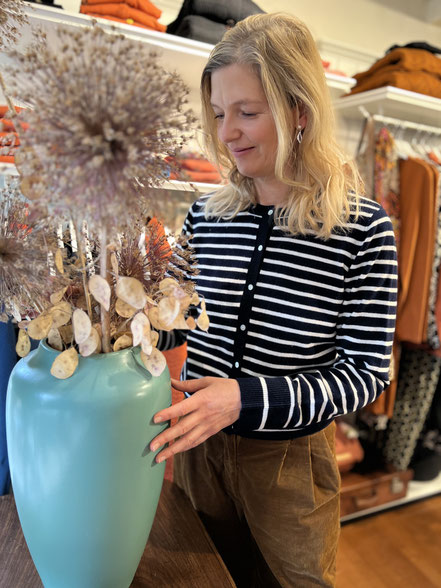 Sonja arrangiert Trockenblumen in türkisfarbener Vase in der Boutique Vergissmeinnicht in Miesbach mit nachhaltiger Mode im Hintergrund.
