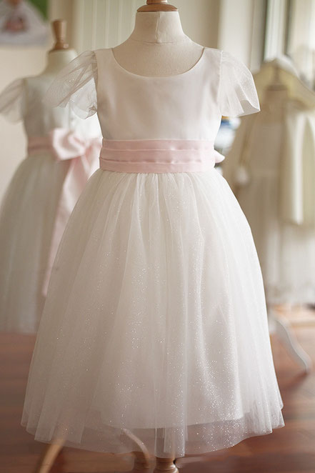 Robe de cérémonie fille en satin blanc cassé et tulle pailleté blanc cassé avec une ceinture rose. Modèle de robe Paola créée par Fil de Légende. Magasin vêtements cérémonie enfants Paris, Neuilly-sur-Seine. Expédition en France et à l'International.