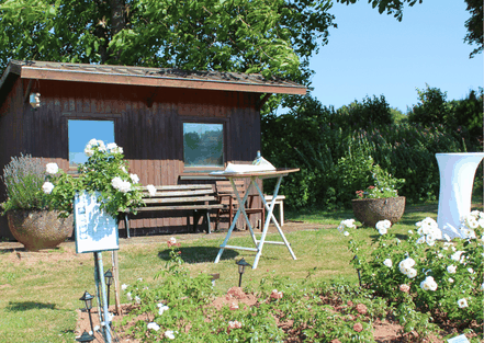 Im Rosengarten Welschbach kann man Familienfeste feiern, hier ist ein Stehtisch vor der Blockhütte zu sehen und ein Rosenbeet
