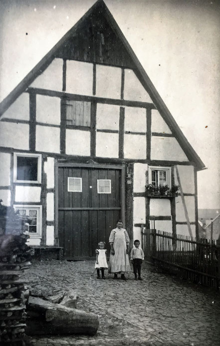 Anna Nolte mit ihren beiden Kindern Theodor und Maria vor ihrem Haus in Freienohl. Am Haus ist die Nummer 47 und rechts weitere Häuser im Dorf zu erkennen. 