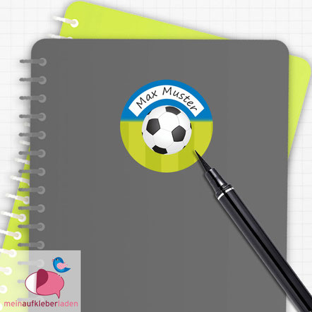 Runder Schulaufkleber mit Fußball-Motiv und dem Namen Max Muster auf einem grauen Schulheft mit Stift.