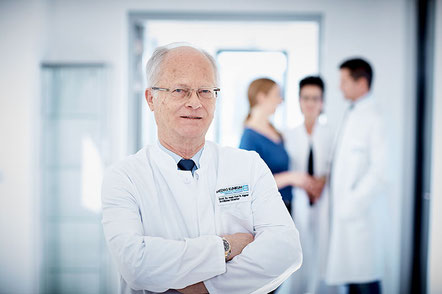 Arzt im weißen Kittel, mit verschränkten Armen, steht freundlich blickend im Vordergrund eines modernen Klinikflurs; im Hintergrund sprechen weitere Ärzte miteinander.