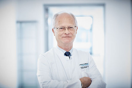 Herr Prof. Dr. med. Karl R. Aigner, Ärztlicher Direktor und Chefarzt, im weißen Kittel mit Brille, steht in einem modernen Klinikflur und schaut freundlich in die Kamera.