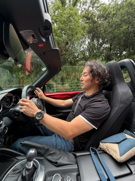 ロードスターのオープンカーを運転する黒いポロシャツを着た男性がオープンカーのロードスターを運転し、その横には帆布バッグのお店Cargo Ship のショルダーバッグcalamelのベージュとブルーのバッグが置いています