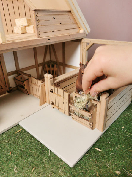 Eine Hand befüllt die Heuraufe an einem Holz-Pferdestall im Schleich® Maßstab 1:24 mit Heu für ein Spielzeugpferd