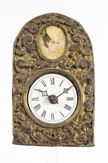 kleine hölzerne Uhr mit einem Uhrenschild aus geprägtem Blech. Das Zifferblatt ist aus weißem Emaile. In der Bekrönung ein Frauenportrait.