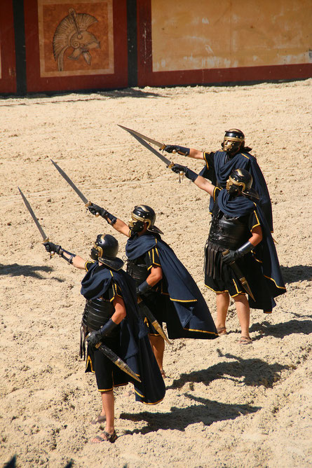 Four men in Roman-style armor and helmets raise swords during a historical reenactment in a sandy arena.