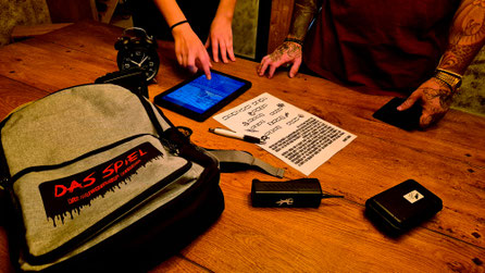 Gemeinsam rätselt die Gruppe beim Escape Spiel. Diverse Requisiten sind auf dem Tisch. Das Spiel kann zu Hause oder an einem Partner Standort selbstständig und ohne persönliche Betreuung gespielt werden.