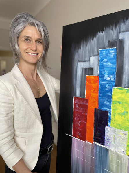 Sara Kouyoumdjian (Barba), artiste peintre, pose tout sourire avec sa toile "au-delà de l'océan". Peinture colorée avec un fond noir. Buildings représentés dans un style semi-figuratif avec plein de couleurs. 