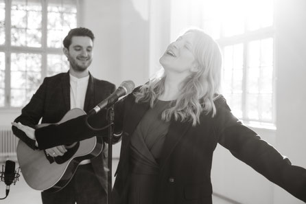 eine Sängerin singt, dahinter der Musiker mit seiner Gitarre lächelt