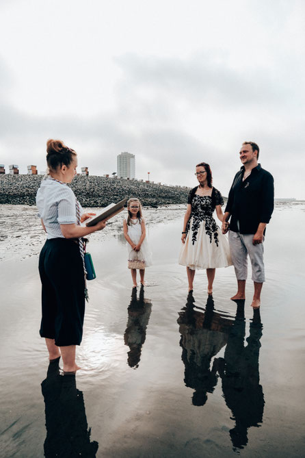 Eine Trauung mitten im Watt, das Brautpaar, die gemeinsame Tochter und Constanze Marienfeld stehen in Büsum mit den Füßen im Wasser. Sie Spiegeln sich im Wasser.