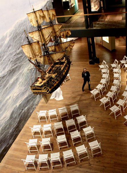 Constanze Marienfeld strahlt in die Kamera im Maritim Museum, über ihr hängt ein Segelschiff, neben ihr sind schon die Stühle für die Trauung aufgebaut.